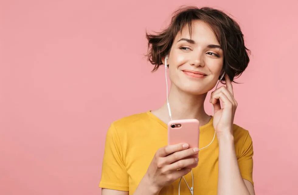 Image of a happy young beautiful woman posing isolated over pink wall background listening music with earphones using mobile phone.