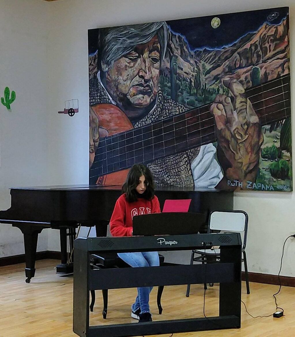 Desde el lienzo, la inspiradora compañía del maestro Ricardo Vilca guía el ensayo de una alumna de la Escuela Superior de Música de Jujuy, que necesita adquirir un nuevo piano.
