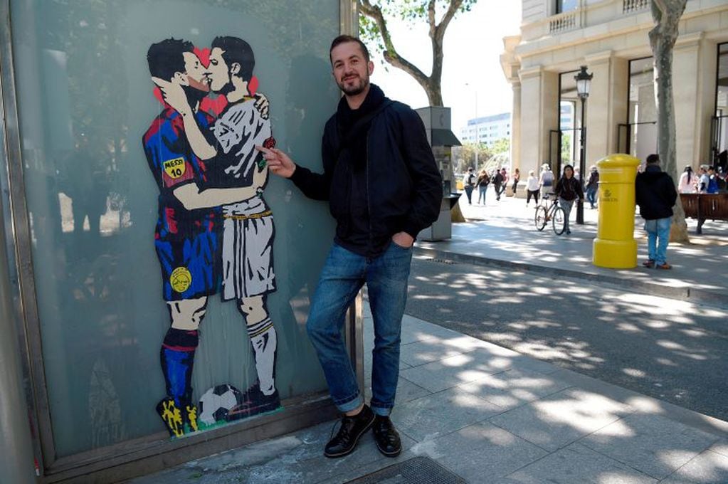Street artist Salva "Tvboy" poses on April 21, 2017 in Barcelona next to his work "Love is blind", which shows Barcelona's Argentinian forward Lionel Messi and Real Madrid's Portuguese forward Cristiano Ronaldo kissing each other two days before 'El Clási