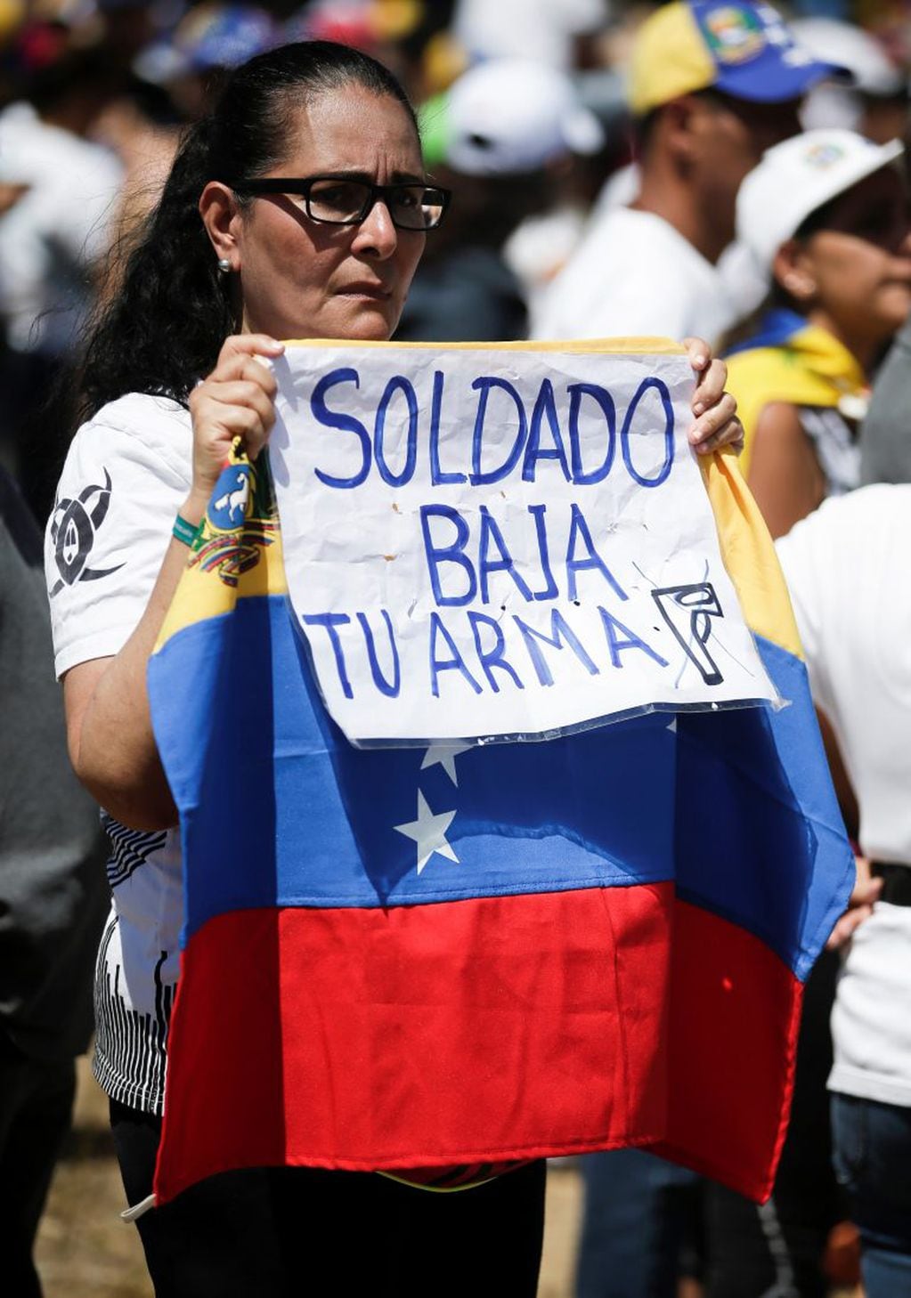 Caracas, Venezuela - Una mujer sostiene una bandera nacional venezolana y un letrero con un mensaje que dice en español: "Soldado baja tu arma" frente a la base militar de La Carlota instando a los soldados a unirse a su lucha y permitir la entrada de ayuda humanitaria de los EE. UU.  (AP Photo/Eduardo Verdugo)