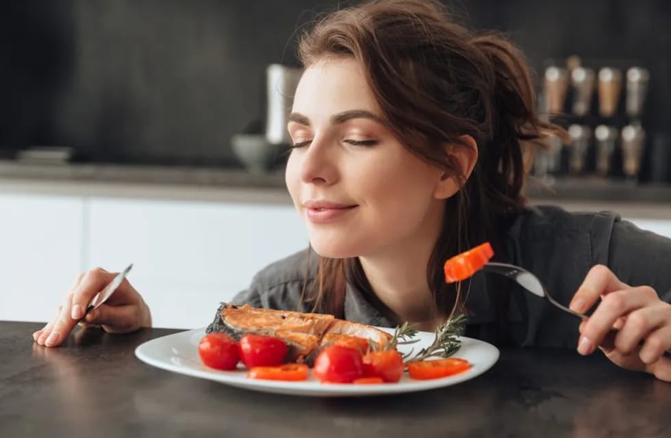 Image of pretty young woman sitting in kitchen while eating and smells fish and tomatoes. Eyes closed.