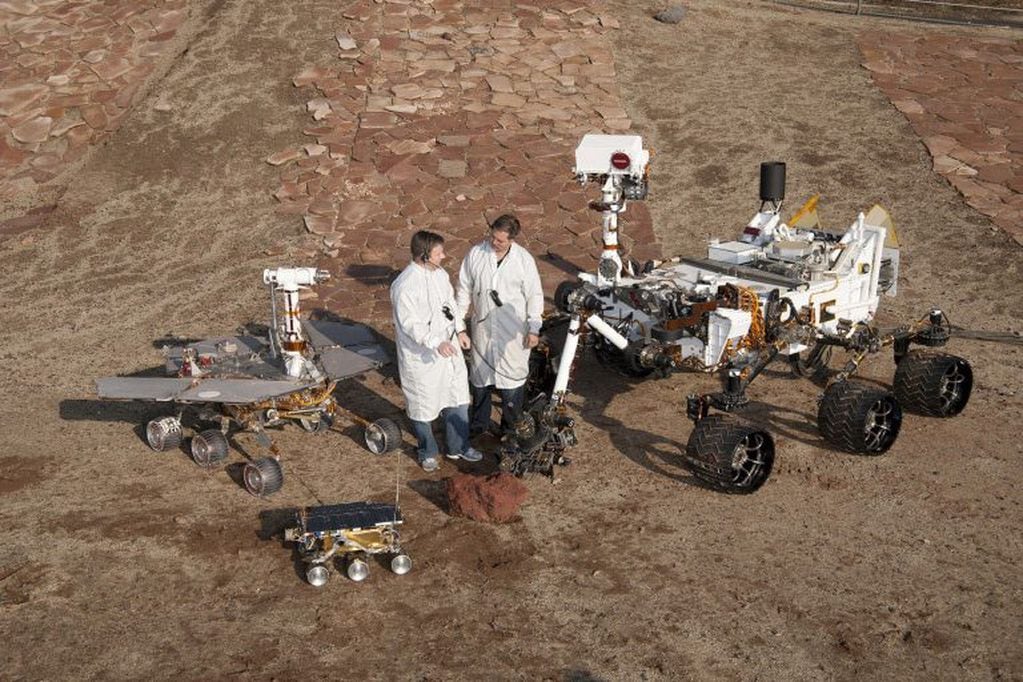 Científicos de la NASA, junto a modelos en tamaño real de las tres generaciones de robots con ruedas enviados ya a Marte. En el extremo izquierdo, el que representa tanto al "Spirit" como al "Opportunity" (2004). Adelante de ellos, el pequeño "Sojourner" (1997). Y a la derecha, el "Curiosity" (2012), de tamaño similar al "Perseverance".