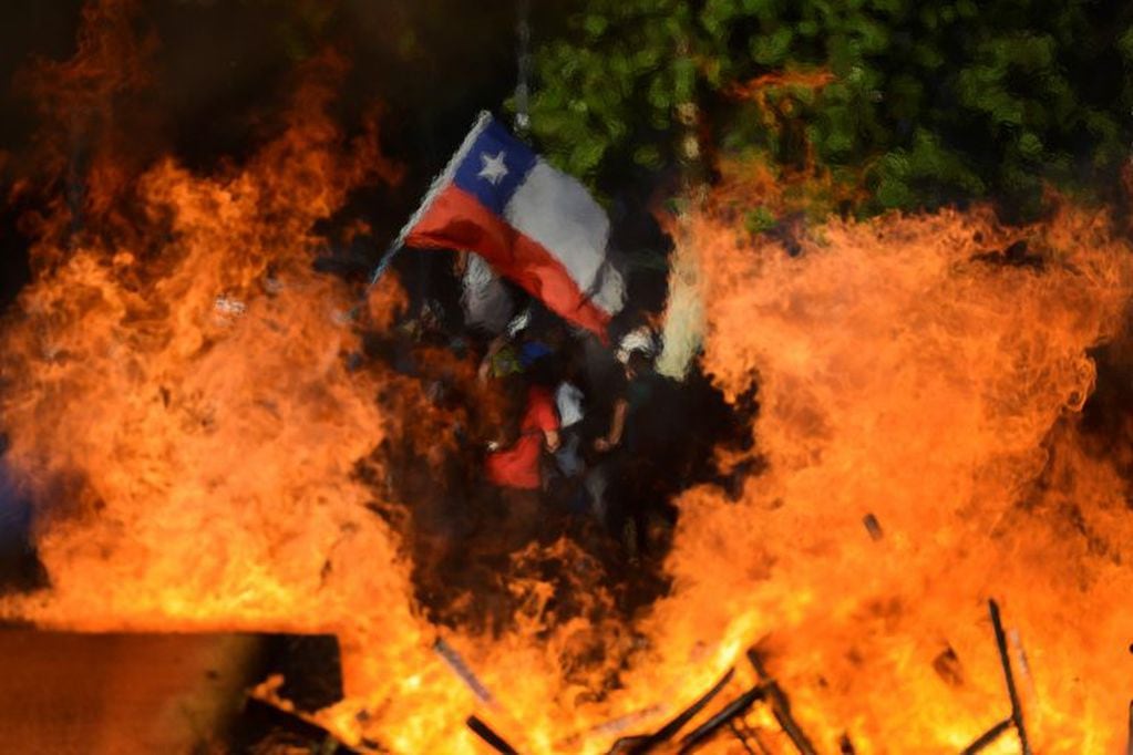 Seen through a burning street barricade, an anti-government demonstrator waves a Chilean flag in Santiago, Chile, Monday, Oct. 28, 2019. Fresh protests and attacks on businesses erupted in Chile Monday despite President Sebastián Piñera's replacement of eight important Cabinet ministers with more centrist figures, and his attempts to assure the country that he had heard calls for greater equality and improved social services. (AP Photo/Matias Delacroix)