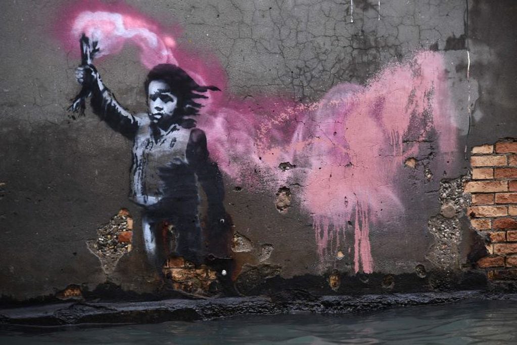 La obra de arte del artista callejero Banksy, que retrata a un niño migrante con un chaleco salvavidas y una bengala rosa neón, se muestra después de un excepcional nivel de agua durante la marea alta. Crédito: Marco Bertorello / AFP.
