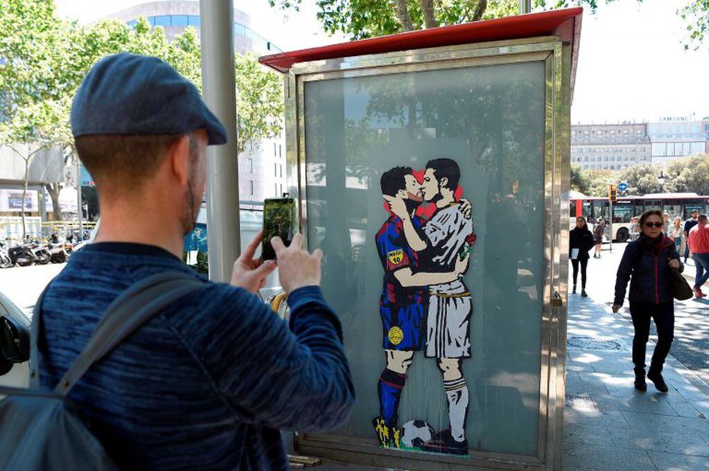 A man takes a picture to the street art "Love is blind" of Street artist Salva "Tvboy" on April 21, 2017 in Barcelona, which shows Barcelona's Argentinian forward Lionel Messi and Real Madrid's Portuguese forward Cristiano Ronaldo kissing each other two d