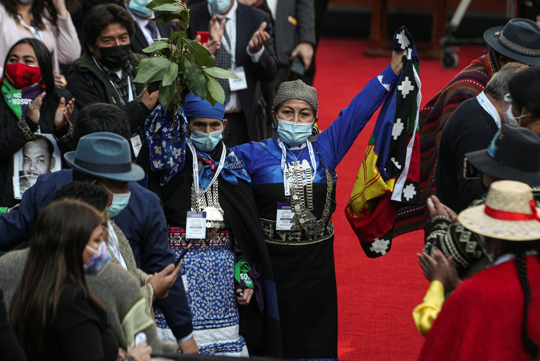 Elisa Loncon, centro, iza una bandera mapuche mientras abraza a su compañera representante Francisca Linconao, una "Machi", después de que Loncon fuera elegida presidenta de la Asamblea Constituyente durante la sesión inaugural de la Convención Constitucional en el edificio del Congreso en Santiago de Chile.