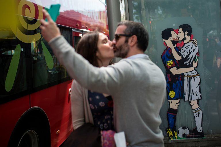 A couple kiss as they take a selfie next to the piece of street art titled "Love is blind", depicting Barcelona's Argentinian forward Lionel Messi (2ndR) and Real Madrid's Portuguese forward Cristiano Ronaldo kissing by street artist Salva "Tvboy", in Bar